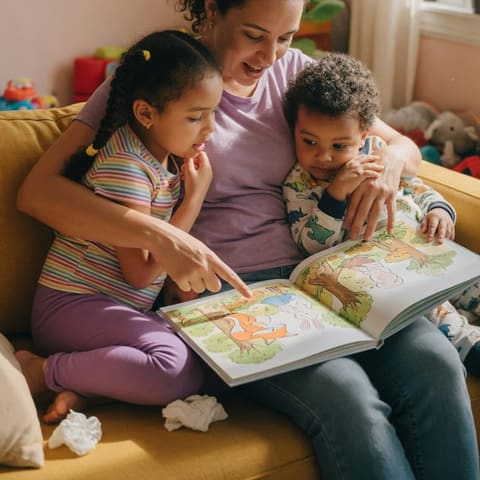 A parent and two young siblings sharing a picture book together on a couch in warm afternoon light